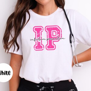 Puede incluir: Camiseta blanca con un gr&aacute;fico rosa "IP" y las palabras "infection prevention" en una fuente cursiva. Un estetoscopio cuelga del cuello. La camiseta es blanca.
