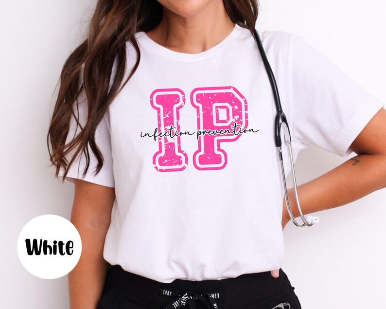 Puede incluir: Camiseta blanca con un gr&aacute;fico rosa "IP" y las palabras "infection prevention" en una fuente cursiva. Un estetoscopio cuelga del cuello. La camiseta es blanca.