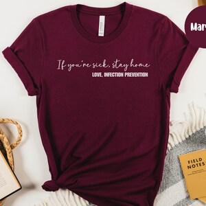 Puede incluir: Camiseta color borgo&ntilde;a con el texto blanco: "If you're sick, stay home. Love, Infection Prevention". La camiseta es de color liso y tiene cuello redondo. La imagen incluye un fondo neutro y una muestra de color etiquetada como "Maroon".