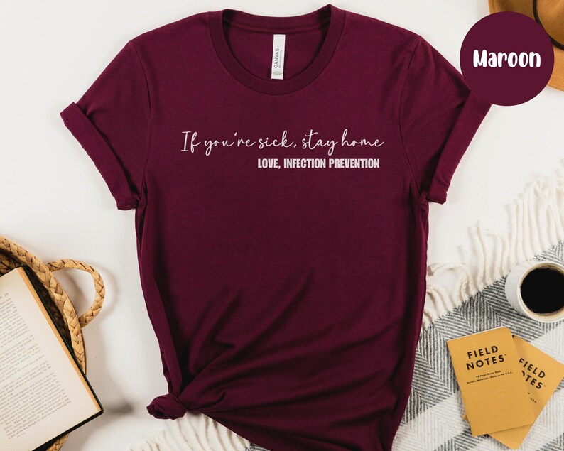 Puede incluir: Camiseta color borgo&ntilde;a con el texto blanco: "If you're sick, stay home. Love, Infection Prevention". La camiseta es de color liso y tiene cuello redondo. La imagen incluye un fondo neutro y una muestra de color etiquetada como "Maroon".