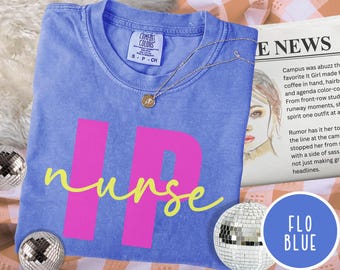 May include: A blue t-shirt with the word "nurse" in yellow script and the letters "HP" in pink. A silver necklace with a small pendant rests on the shirt. The shirt is labeled "Comfort Colors" and "S-P-CH".