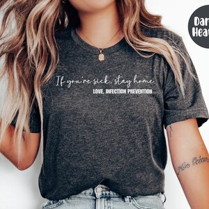 Puede incluir: Camiseta gris jaspeado oscuro con el texto "If you're sick, stay home. Love, Infection Prevention" en escritura blanca. La camiseta tiene cuello redondo y mangas cortas. Un c&iacute;rculo gris oscuro con las palabras "Dark 6. Heather" est&aacute; en la esquina superior derecha.