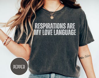 Puede incluir: Camiseta gris oscuro con el texto blanco "RESPIRATIONS ARE MY LOVE LANGUAGE" en letras desgastadas. La camiseta tiene cuello redondo y mangas cortas. Una etiqueta redonda en la esquina inferior izquierda dice "PEPPER".