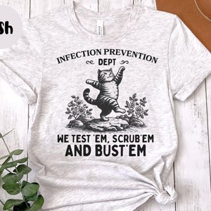 May include: Ash-colored t-shirt featuring a whimsical illustration of a cat balancing on a rock, with the text "INFECTION PREVENTION DEPT" above and "WE TEST 'EM, SCRUB 'EM AND BUST 'EM" below. The cat is in a playful pose.