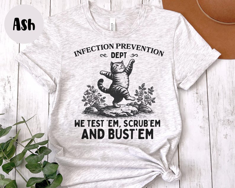 May include: Ash-colored t-shirt featuring a whimsical illustration of a cat balancing on a rock, with the text "INFECTION PREVENTION DEPT" above and "WE TEST 'EM, SCRUB 'EM AND BUST 'EM" below. The cat is in a playful pose.