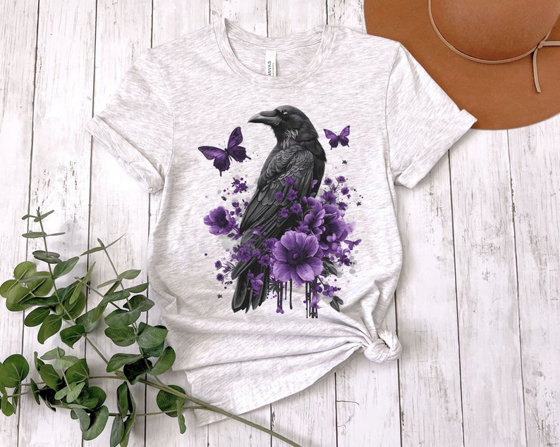 Może przedstawiać: Biały t-shirt z nadrukiem czarnego kruka i fioletowych kwiat&oacute;w z przodu. Kruk siedzi na gałęzi z rozpostartymi skrzydłami. Kwiaty są w rozkwicie i mają efekt akwarelowy.