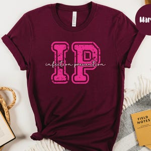 Puede incluir: Camiseta color granate con un gr&aacute;fico "IP" rosa desgastado y la frase "infection prevention" en cursiva. La camiseta es de material suave y tiene las mangas dobladas.