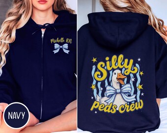 Puede incluir: Una sudadera con capucha y cremallera azul marino que presenta un gráfico de lazo a rayas blancas y azul claro con el texto Michelle RN en el frente. La parte posterior muestra un gráfico de un pato con un lazo a rayas, el texto Silly Peds crew y estrellas amarillas.