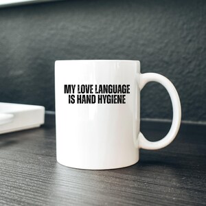 May include: White ceramic mug with a curved handle. The mug features the text "MY LOVE LANGUAGE IS HAND HYGIENE" in bold black letters. The mug is on a dark wooden surface.