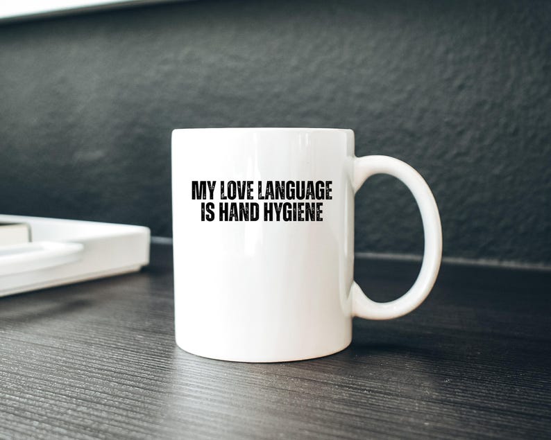 May include: White ceramic mug with a curved handle. The mug features the text "MY LOVE LANGUAGE IS HAND HYGIENE" in bold black letters. The mug is on a dark wooden surface.