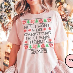 May include: A light pink t-shirt with a Christmas-themed design. The design includes the text "ALL I WANT FOR CHRISTMAS IS CLEAN HANDS 2025" in a pixelated font, along with red snowflakes and green Christmas trees.