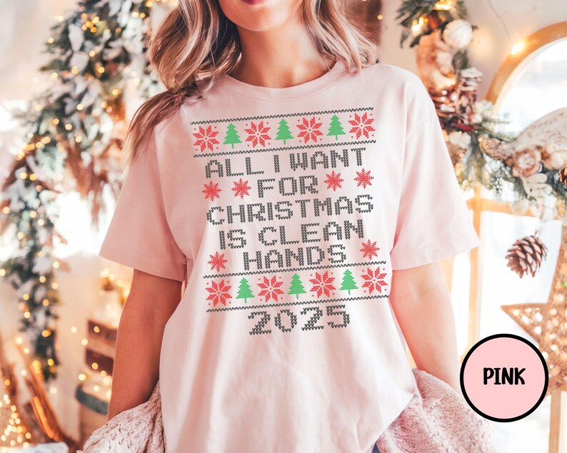 May include: A light pink t-shirt with a Christmas-themed design. The design includes the text "ALL I WANT FOR CHRISTMAS IS CLEAN HANDS 2025" in a pixelated font, along with red snowflakes and green Christmas trees.