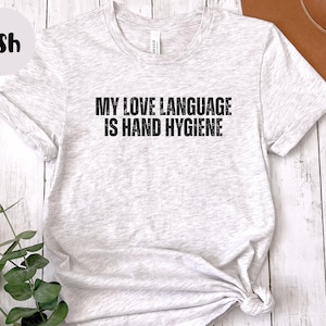 May include: Light gray t-shirt with the text "MY LOVE LANGUAGE IS HAND HYGIENE" in black. The shirt is knotted at the bottom and has rolled-up sleeves. The word "Ash" is in a circle in the upper left corner.