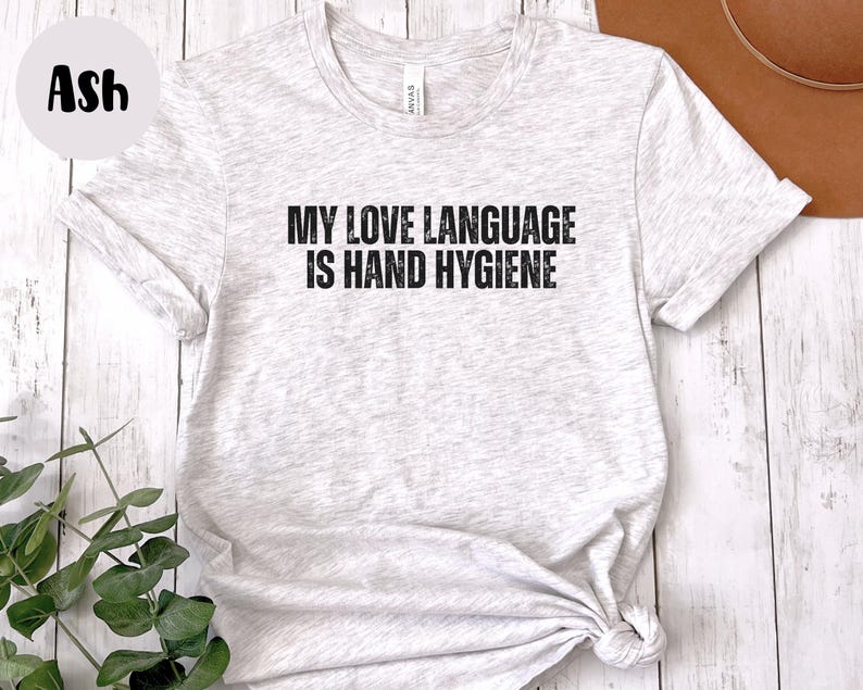 May include: Light gray t-shirt with the text "MY LOVE LANGUAGE IS HAND HYGIENE" in black. The shirt is knotted at the bottom and has rolled-up sleeves. The word "Ash" is in a circle in the upper left corner.