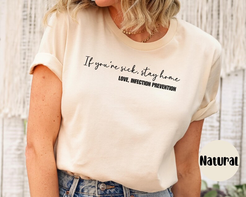 Puede incluir: Una camiseta de color natural con el texto "If you're sick, stay home" en escritura negra. Debajo, "LOVE, INFECTION PREVENTION" en negrita negra. La camiseta tiene mangas enrolladas.