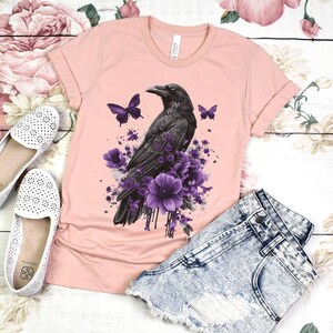 Może przedstawiać: R&oacute;żowy T-shirt z nadrukiem czarnego kruka i fioletowych kwiat&oacute;w z przodu. Kruk jest otoczony fioletowymi kwiatami i motylami. Koszulka leży na białym tle z parą białych but&oacute;w i dżinsowymi spodenkami.