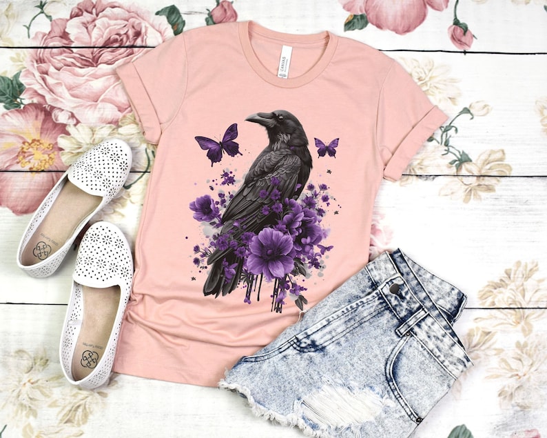 Może przedstawiać: R&oacute;żowy T-shirt z nadrukiem czarnego kruka i fioletowych kwiat&oacute;w z przodu. Kruk jest otoczony fioletowymi kwiatami i motylami. Koszulka leży na białym tle z parą białych but&oacute;w i dżinsowymi spodenkami.