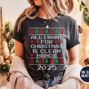 May include: Dark grey heather t-shirt with a festive Christmas sweater design. The shirt features the text "ALL I WANT FOR CHRISTMAS IS CLEAN HANDS 2025" in white, red, and green. The design includes Christmas trees and snowflake patterns.