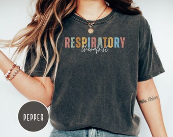 Puede incluir: Camiseta gris oscuro con las palabras "RESPIRATORY therapist" en letras coloridas de estilo vintage. La camiseta tiene cuello redondo y mangas cortas. La palabra "Pepper" está en un círculo en la parte inferior izquierda.