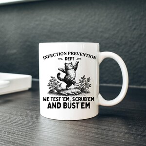 May include: White ceramic mug with a black and white illustration of a cat standing on its hind legs. The mug has the text "INFECTION PREVENTION DEPT" and "WE TEST 'EM, SCRUB 'EM AND BUST 'EM".