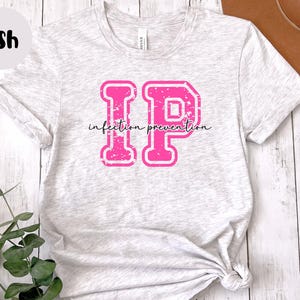 Puede incluir: Camiseta gris ceniza con un gr&aacute;fico "IP" rosa, con las palabras "infection prevention" en una fuente cursiva. Las letras tienen un aspecto desgastado y vintage. La camiseta est&aacute; hecha de un material suave y claro.