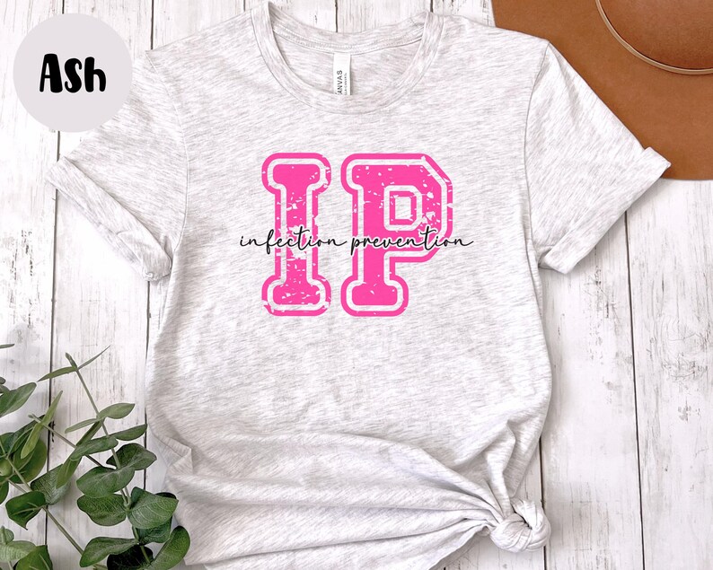 Puede incluir: Camiseta gris ceniza con un gr&aacute;fico "IP" rosa, con las palabras "infection prevention" en una fuente cursiva. Las letras tienen un aspecto desgastado y vintage. La camiseta est&aacute; hecha de un material suave y claro.