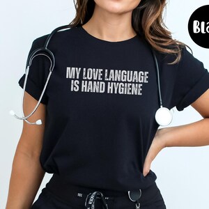 May include: Black t-shirt with the text "MY LOVE LANGUAGE IS HAND HYGIENE" in white, distressed lettering. A stethoscope is draped around the neck. The shirt is a crew neck style.