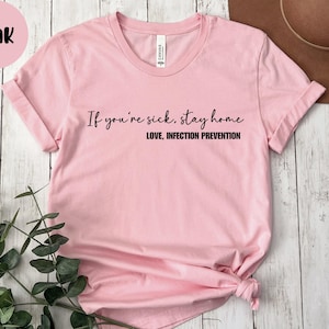 Puede incluir: Camiseta rosa claro con el texto negro "If you're sick, stay home. Love, Infection Prevention". La camiseta tiene cuello redondo y mangas cortas. Un c&iacute;rculo rosa con la palabra "Pink" est&aacute; en la esquina superior izquierda.