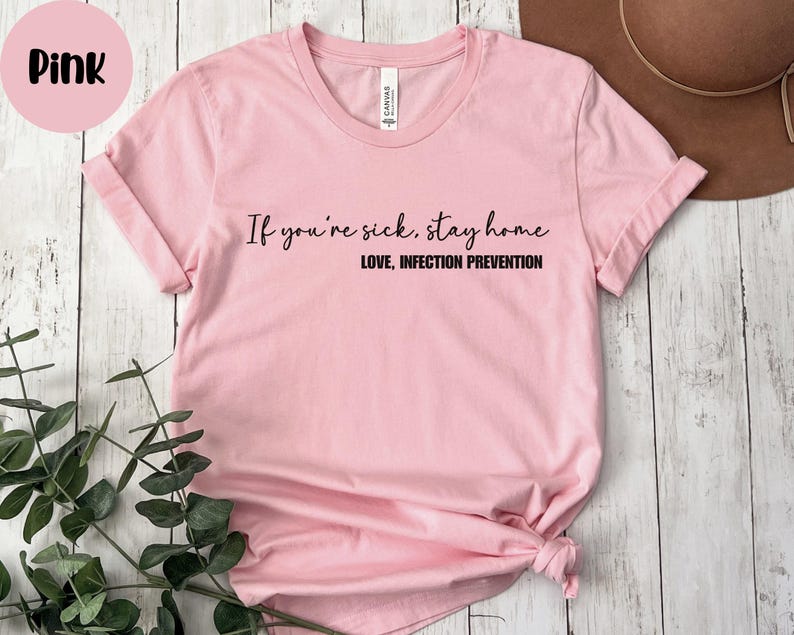 Puede incluir: Camiseta rosa claro con el texto negro "If you're sick, stay home. Love, Infection Prevention". La camiseta tiene cuello redondo y mangas cortas. Un c&iacute;rculo rosa con la palabra "Pink" est&aacute; en la esquina superior izquierda.