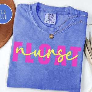 May include: A blue Comfort Colors t-shirt with the word "FLOAT" in pink block letters and "nurse" in yellow cursive. A gold necklace with a pendant is draped across the shirt. The shirt is folded and displayed on a white surface.
