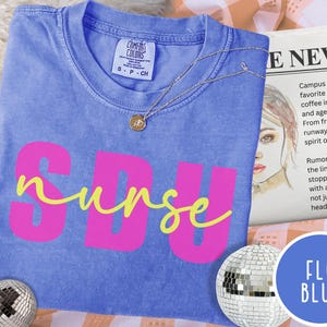 May include: A blue t-shirt with the word "SBU" in pink block letters and the word "nurse" in yellow script. A silver necklace with a pendant is on the shirt. The shirt is folded on a patterned surface.