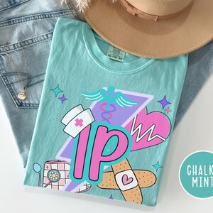 May include: A light teal t-shirt with a graphic design featuring medical symbols, a nurse's hat, and the letters "IP" in pink. The shirt is folded on top of blue jeans and a tan hat. The words "Chalky Mint" are in a circle.