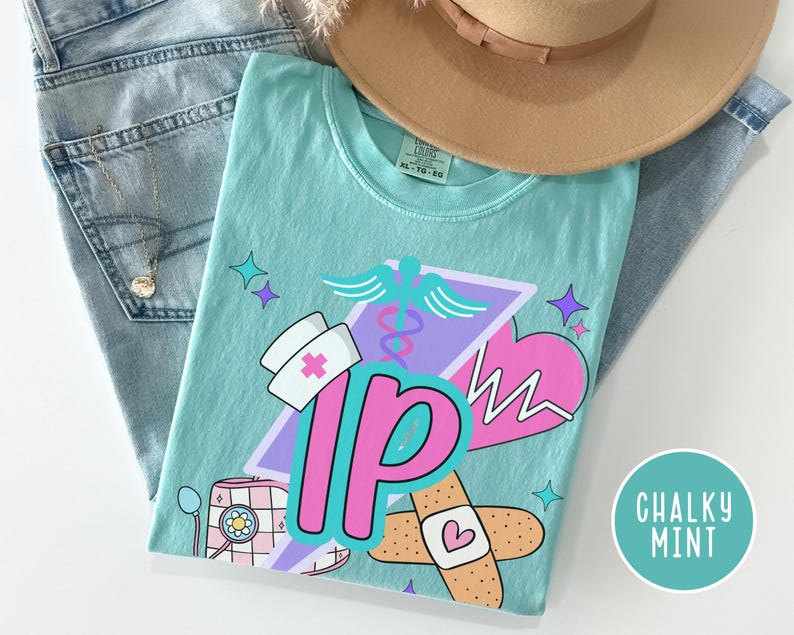 May include: A light teal t-shirt with a graphic design featuring medical symbols, a nurse's hat, and the letters "IP" in pink. The shirt is folded on top of blue jeans and a tan hat. The words "Chalky Mint" are in a circle.