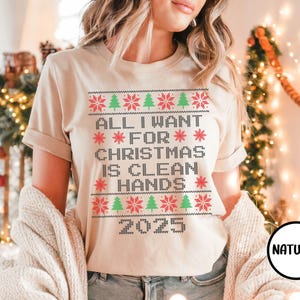 May include: A beige t-shirt with a Christmas-themed design. The text reads "ALL I WANT FOR CHRISTMAS IS CLEAN HANDS 2025" in a pixelated font, with red snowflakes and green Christmas trees. The shirt has short sleeves.