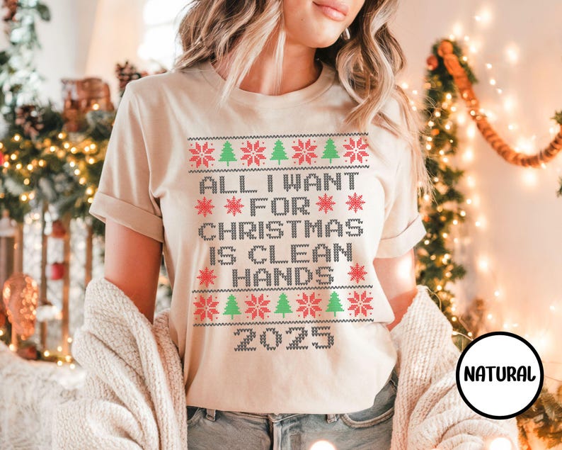May include: A beige t-shirt with a Christmas-themed design. The text reads "ALL I WANT FOR CHRISTMAS IS CLEAN HANDS 2025" in a pixelated font, with red snowflakes and green Christmas trees. The shirt has short sleeves.