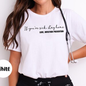 Puede incluir: Camiseta blanca con el texto "If you're sick, stay home. Love, Infection Prevention" en negro. Un estetoscopio cuelga del cuello. La palabra "White" est&aacute; en un c&iacute;rculo blanco en la esquina inferior izquierda.