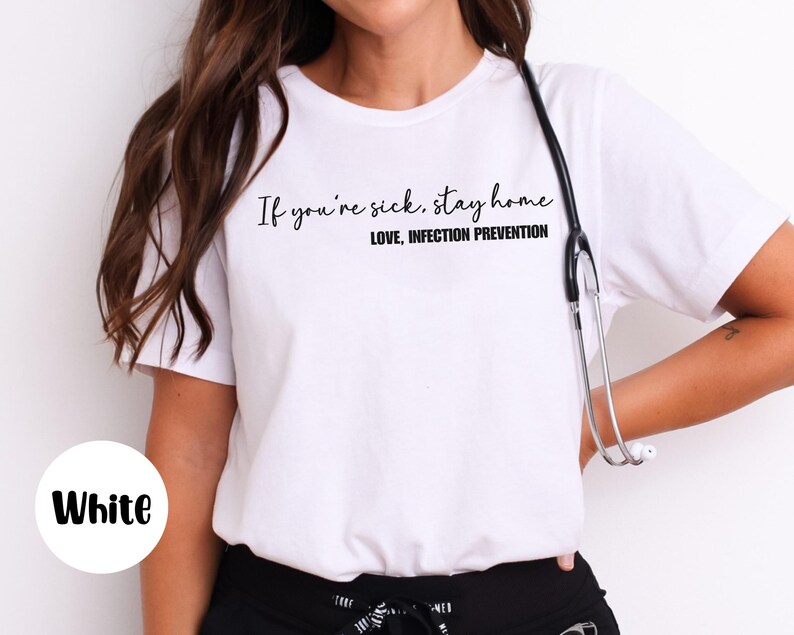 Puede incluir: Camiseta blanca con el texto "If you're sick, stay home. Love, Infection Prevention" en negro. Un estetoscopio cuelga del cuello. La palabra "White" est&aacute; en un c&iacute;rculo blanco en la esquina inferior izquierda.