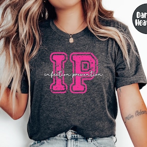 Puede incluir: Camiseta gris oscuro jaspeado con un gr&aacute;fico "IP" rosa desgastado y las palabras "infection prevention" en fuente cursiva. La camiseta es de cuello redondo.