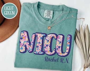 Puede incluir: Camiseta verde claro con la palabra "NICU" en letras grandes llenas de flores con un contorno morado. El nombre "Rachel R.N." está impreso debajo. Un collar de oro con un pequeño colgante descansa sobre la camisa.