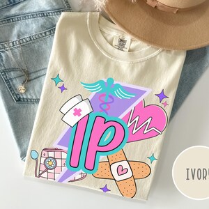 May include: Ivory-colored t-shirt with a colorful graphic design featuring medical symbols, a nurse's hat, and the letters "IP". The design includes a caduceus, a heart monitor, and a bandage. The shirt is folded on top of blue jeans.