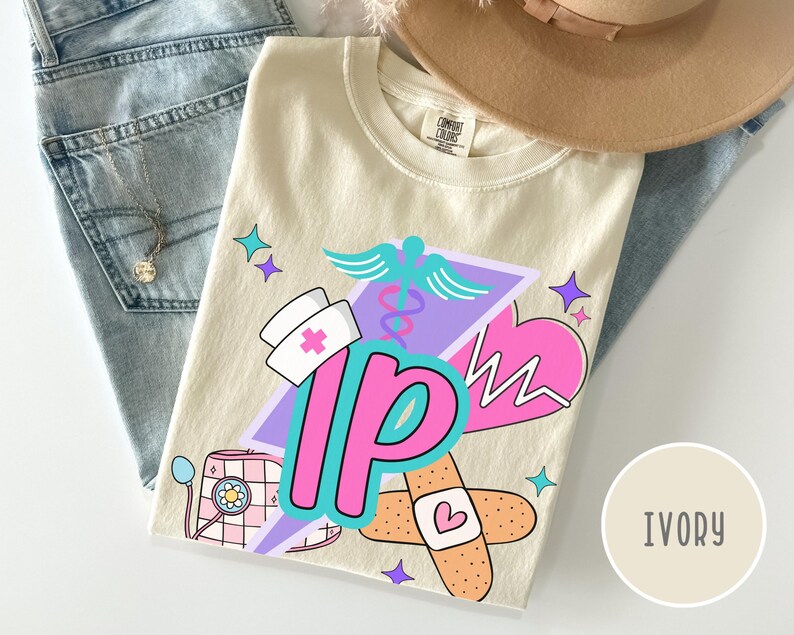 May include: Ivory-colored t-shirt with a colorful graphic design featuring medical symbols, a nurse's hat, and the letters "IP". The design includes a caduceus, a heart monitor, and a bandage. The shirt is folded on top of blue jeans.