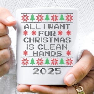 May include: White ceramic mug with a festive design. The mug features the text "ALL I WANT FOR CHRISTMAS IS CLEAN HANDS 2025" in a pixelated font, with red snowflakes and green Christmas trees. The mug is held by a person.