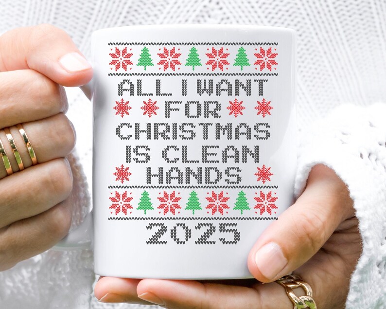 May include: White ceramic mug with a festive design. The mug features the text "ALL I WANT FOR CHRISTMAS IS CLEAN HANDS 2025" in a pixelated font, with red snowflakes and green Christmas trees. The mug is held by a person.