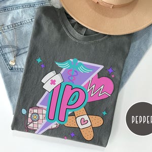 May include: A gray t-shirt featuring a colorful graphic design with a nurse's hat, a heart, and the letters "IP". The design includes a caduceus, a bandage, and stars. A tan hat and blue jeans are also in the image.