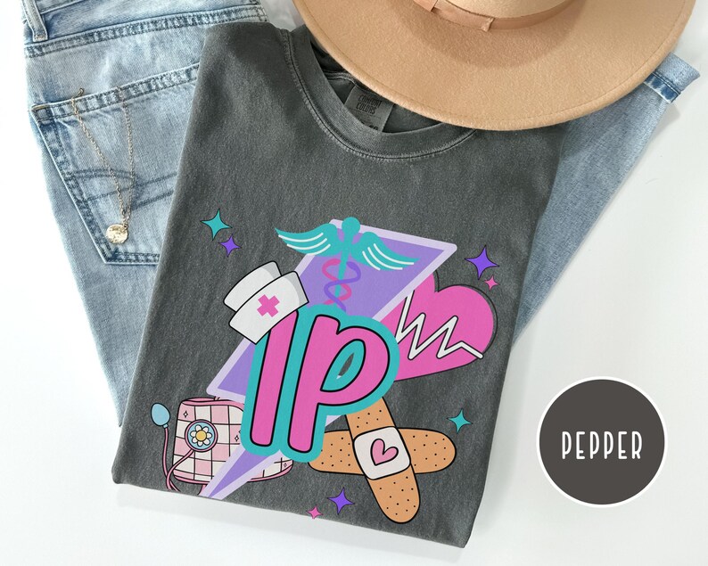 May include: A gray t-shirt featuring a colorful graphic design with a nurse's hat, a heart, and the letters "IP". The design includes a caduceus, a bandage, and stars. A tan hat and blue jeans are also in the image.