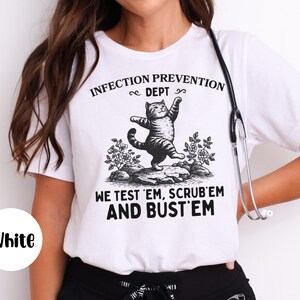 May include: White t-shirt with a black and white graphic of a cat standing on a rock with the text "INFECTION PREVENTION DEPT" and "WE TEST 'EM, SCRUB 'EM AND BUST 'EM". A stethoscope hangs on the shirt.
