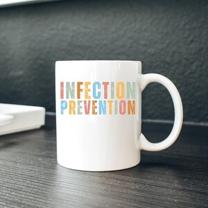 May include: White ceramic mug with the words "INFECTION PREVENTION" in colorful, vintage-style lettering. The mug has a curved handle and sits on a dark wooden surface. The background is a dark gray.