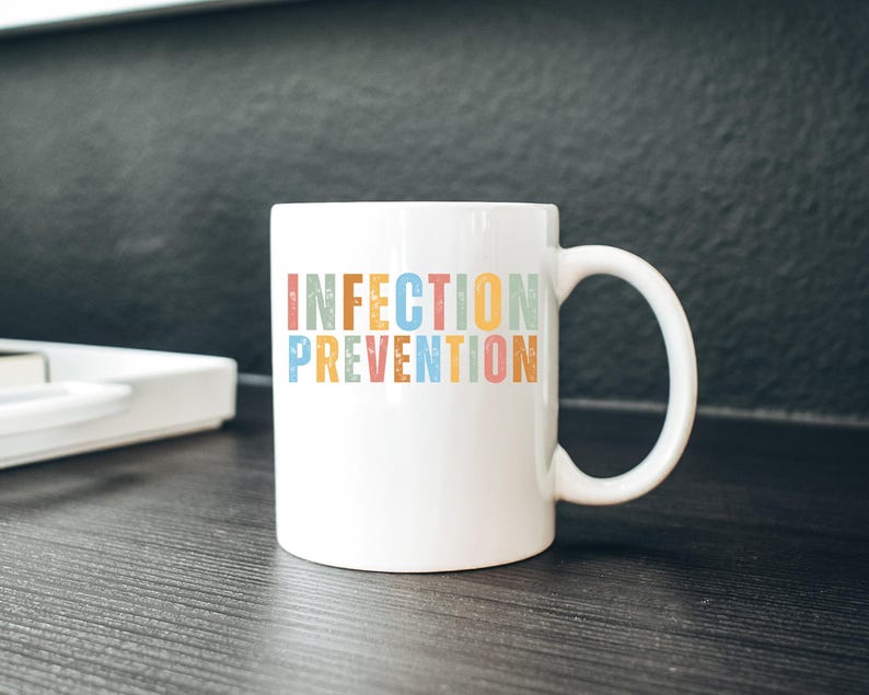 May include: White ceramic mug with the words "INFECTION PREVENTION" in colorful, vintage-style lettering. The mug has a curved handle and sits on a dark wooden surface. The background is a dark gray.