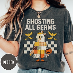 May include: Dark gray t-shirt with the text "Ghosting All Germs" above a cartoon ghost wearing a patchwork quilt and holding a jack-o'-lantern. The design includes bats and a checkered pattern, perfect for Halloween.