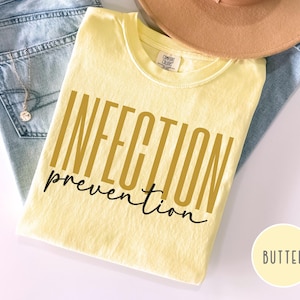 May include: A light yellow t-shirt with the words "INFECTION prevention" printed in gold and black. The shirt is folded and placed on top of blue jeans and a tan hat. A small circle with the word "BUTTER" is in the bottom right corner.